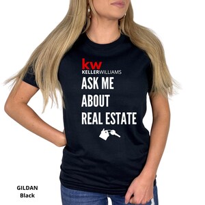 May include: Black t-shirt with the Keller Williams logo in red and the text "ASK ME ABOUT REAL ESTATE" in white. A graphic of a house and key is below the text. The shirt is a Gildan brand.