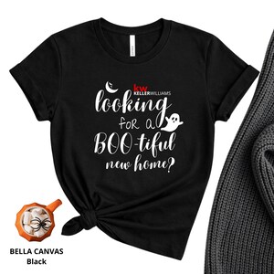 May include: A black short-sleeved t-shirt featuring the white text "looking for a BOO-tiful new home?" with a small ghost and crescent moon graphic, plus the red and white "KW KELLERWILLIAMS" logo. The shirt is knotted at the bottom hem.