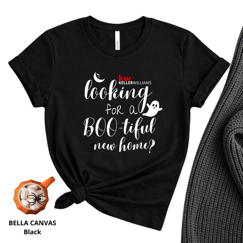 May include: A black short-sleeved t-shirt featuring the white text "looking for a BOO-tiful new home?" with a small ghost and crescent moon graphic, plus the red and white "KW KELLERWILLIAMS" logo. The shirt is knotted at the bottom hem.