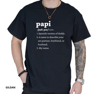 May include: A navy blue t-shirt with the word "papi" in white, followed by a definition. The definition includes the Spanish version of daddy, a name for a partner, and "My name."