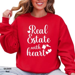 May include: A red sweatshirt with the text "Real Estate with heart" in white script. The design includes a house, key, and heart symbols. The sweatshirt is a Gildan brand.