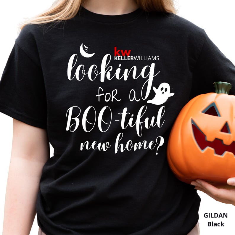 May include: A black short-sleeved t-shirt featuring white text that reads "looking for a BOO-tiful new home?" along with a small white crescent moon, a ghost graphic, and the red and white "kw KELLERWILLIAMS" logo printed on the front.