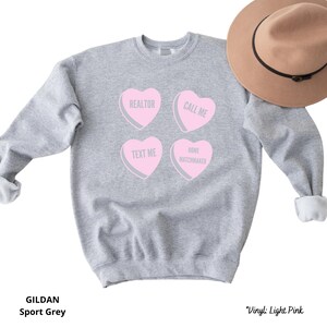 May include: A light gray sweatshirt with four pink heart-shaped graphics. Each heart has a different message: "REALTOR", "CALL ME", "TEXT ME", and "HOME MATCHMAKER". The sweatshirt is a Gildan Sport Grey. A brown hat is in the background.