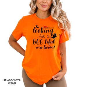 May include: A bright orange short-sleeved t-shirt featuring black text that reads "looking for a BOO-tiful new home?" with a small ghost graphic and the red and black "kw KELLERWILLIAMS" logo printed on the front chest area.