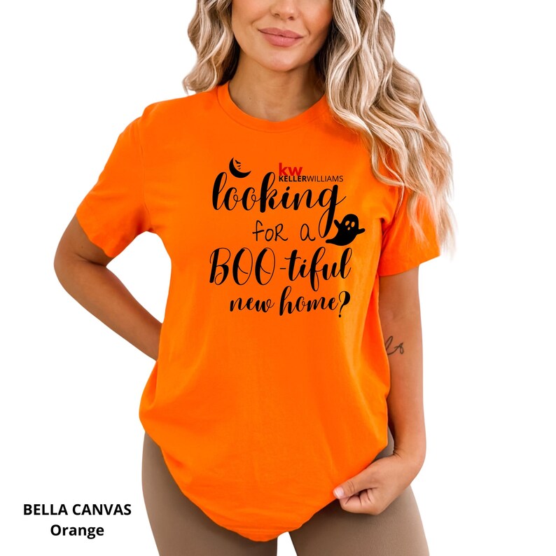 May include: A bright orange short-sleeved t-shirt featuring black text that reads "looking for a BOO-tiful new home?" with a small ghost graphic and the red and black "kw KELLERWILLIAMS" logo printed on the front chest area.