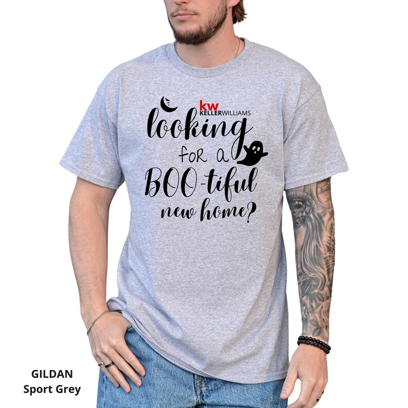 May include: A light grey short-sleeve t-shirt featuring black text that reads "looking for a BOO-tiful new home?" with a small black crescent moon and ghost graphic. The shirt also includes the red and black Keller Williams logo above the text.