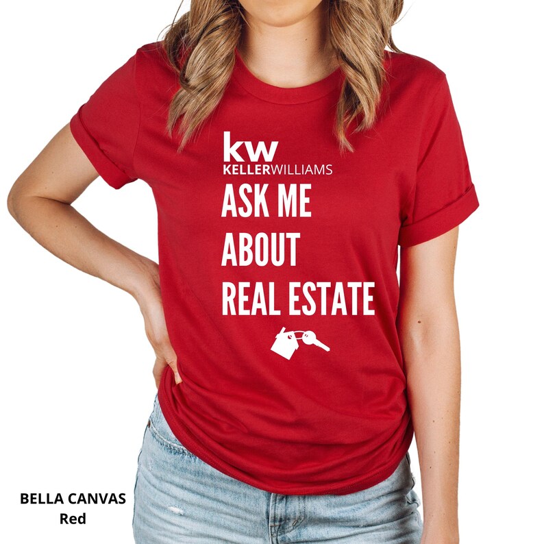 May include: A red Bella Canvas t-shirt with white text that reads "kw KELLER WILLIAMS ASK ME ABOUT REAL ESTATE" with a house and key graphic. The shirt has short sleeves and is being worn.