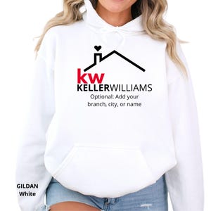 May include: A white hooded sweatshirt featuring a black and red graphic logo of a house roof with a heart above the chimney. Below the roof, the text reads kw KELLERWILLIAMS and Optional: Add your branch, city, or name. The brand is Gildan.