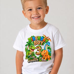 Könnte beinhalten: Ein weißes kurzärmeliges T-Shirt mit einer bunten Grafik aus drei Dinosauriern, Palmen und dem Text "BIRTHDAY BOY" über einer großen Zahl "3" in Steinoptik. Der Name "JOSHUA" ist unten in schwarzen Buchstaben aufgedruckt.