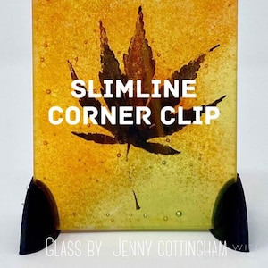 May include: A square piece of glass with a yellow, orange, and green gradient. A black silhouette of a leaf is in the centre of the glass. The text "SLIMLINE CORNER CLIP" is printed in white on the glass. The glass is sitting on two black corner clips.
