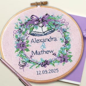 May include: A cross-stitch embroidery hoop featuring a floral wreath design in shades of purple, blue, and green, surrounding the names "Alexandra & Mathew" and the date "12.09.2025". The hoop is set against a light pink fabric.