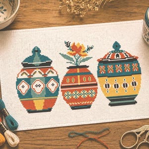 Può includere: Un disegno a punto croce con tre vasi decorativi con coperchi, ognuno decorato con motivi geometrici in verde acqua, arancione, giallo e nero. I vasi sono su uno sfondo bianco, con filo da ricamo e forbici nelle vicinanze. Il disegno è su una superficie di legno.