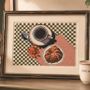 Pode incluir: A arte em ponto cruz emoldurada retrata uma xícara de café, um croissant e uma flor em um fundo quadriculado. Uma caneca "Café de Paris" e um quadro-negro com preços também são visíveis. A arte é exibida em uma superfície de madeira.