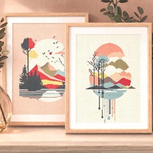 May include: Two framed cross-stitch landscape art pieces. One features a sunset over mountains and trees, the other a similar scene with a different colour palette. Both are in light wood frames, displayed on a light-coloured surface.