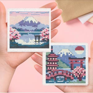 May include: Two small cross-stitch artworks depicting Japanese landscapes. One features Mount Fuji, cherry blossoms, and a torii gate. The other shows a pagoda, bridge, and the rising sun. The artworks are held in hands against a pink background.