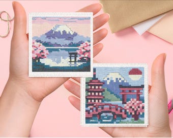 May include: Two square cross-stitch embroidery pieces featuring Japanese landscapes. One depicts a mountain with cherry blossoms and a torii gate. The other shows a pagoda, a red bridge, a torii gate, a mountain, and a sun against a blue sky.