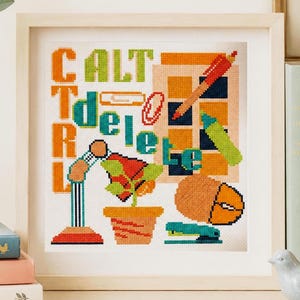 Pode incluir: Obra de arte em ponto cruz emoldurada em madeira com as palavras "CTRL DELETE". O design inclui uma caneta, um marcador, um clipe de papel e outros elementos com tema de escritório. A obra de arte é exibida em uma prateleira com livros.