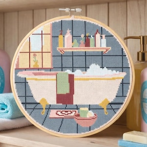 Puede incluir: Un bordado de punto de cruz en un aro de madera representa una escena de baño con una bañera, una estantería con botellas y una ventana. Botellas rosas y azules de loción y jabón están en la estantería. Una toalla azul doblada y una flor rosa están en primer plano.