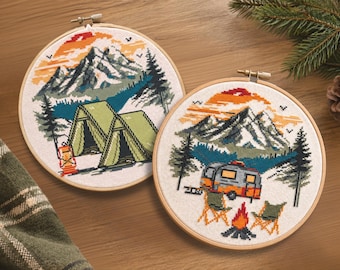 May include: Two circular wooden embroidery hoops containing cross-stitch art. One hoop features a green tent, a lantern, and mountains under an orange sunset. The other hoop displays a camper trailer, two chairs, and a campfire before mountains.