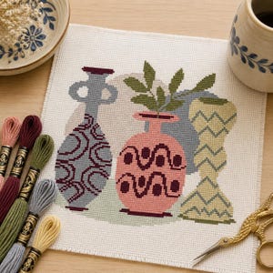 May include: A cross-stitch design depicting three vases in grey, pink, and yellow, with green leaves in one. Embroidery threads in various colours and gold scissors are arranged around the design, with a ceramic bowl and mug.