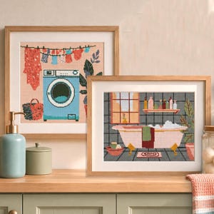 May include: Two framed art prints depicting a laundry room and a bathroom scene. The laundry room print shows a blue washing machine and clothes hanging on a line. The bathroom print features a pink bathtub with bubbles and toiletries.