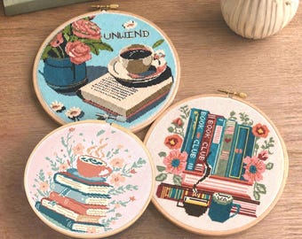 May include: Three circular wooden embroidery hoops featuring cross-stitch designs. One shows a blue background with a book, coffee cup, and pink flowers with the text UNWIND. Another displays a stack of books with a coffee cup and pink flowers.