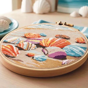 May include: A cross-stitch embroidery hoop depicting a beach scene with vibrant seashells. The design showcases orange, purple, and blue shells on a sandy backdrop, enclosed within a wooden hoop. The image suggests a coastal theme.