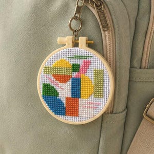 Puede incluir: Un llavero circular de madera con un patrón de punto de cruz de formas geométricas abstractas en amarillo, naranja, azul, verde y rosa sobre un fondo de tela blanca, unido a una anilla y un cierre de metal.