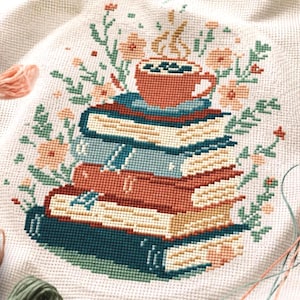 May include: Cross-stitch design featuring a stack of books with a coffee cup on top, surrounded by floral elements. The books are in shades of red, blue, and beige. The cup is orange, and the flowers are peach and green.