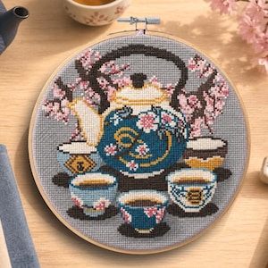 May include: Cross-stitch embroidery of a blue teapot and teacups, surrounded by pink cherry blossoms. The design is set in a wooden embroidery hoop. A blue teapot and tea towels are in the background.