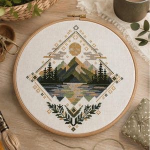 May include: A circular wooden embroidery hoop containing a cross-stitch pattern of a mountain landscape. The design features a sun, clouds, pine trees, and a lake in shades of green, blue, and gold, framed within a diamond-shaped geometric border.