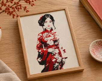 May include: A framed cross-stitch embroidery featuring a person with dark hair styled in an updo, wearing a red and black patterned garment with white floral accents. The artwork is displayed in a light-colored wooden frame on a flat surface.