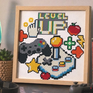 May include: Cross-stitch artwork in a wooden frame with a pixelated gaming theme. The design includes a controller, joystick, and the words "LEVEL UP." The colour palette includes grey, green, red, yellow, and blue.