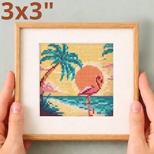 May include: A framed 3x3 inch cross-stitch artwork featuring a flamingo on a beach at sunset. The artwork includes a palm tree, ocean, and a large orange sun. The frame is made of light-colored wood.