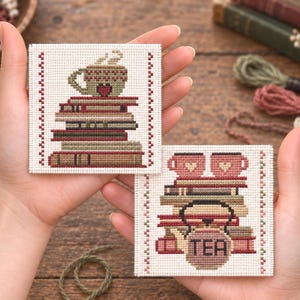 May include: Two square cross-stitch patterns on white fabric. One features a stack of books topped with a steaming mug with a heart. The other shows a stack of books with two mugs and a teapot labelled TEA. Both have red stitched borders.