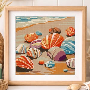 May include: A framed artwork featuring a pixelated beach scene with colorful seashells. The shells are in shades of orange, blue, purple, and white, scattered on a sandy shore. The artwork is in a light wooden frame.