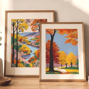 May include: Two framed cross-stitch landscape art pieces. One features a river scene with colorful trees in autumn hues. The other depicts a path through a forest with vibrant orange and yellow foliage. Both are in light wood frames.