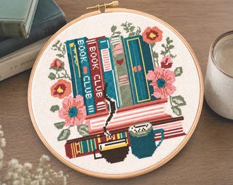 May include: Cross-stitch embroidery in a wooden hoop features a stack of books with the text "BOOK CLUB III", surrounded by pink and orange flowers, and a blue mug with a coffee cup design. The background is a light beige fabric.