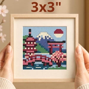 May include: A framed 3x3 inch cross-stitch depicting a Japanese landscape. The artwork features a pagoda, bridge, torii gate, Mount Fuji, and a rising sun. The color palette includes shades of blue, pink, red, and green, all within a light wood frame.