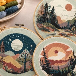 Könnte beinhalten: Drei runde Stickrahmen mit Landschaftsmotiven. Einer zeigt eine Nachtszene mit Mond und Sternen, ein anderer einen Sonnenuntergang und der dritte eine Berglandschaft bei Tag. Stickgarn in verschiedenen Farben befindet sich in einer kleinen Schale.