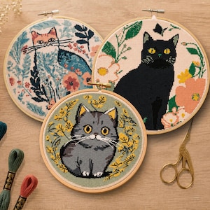 May include: Three circular wooden embroidery hoops containing cross-stitch designs of cats. One features a white cat with blue foliage, another a black cat with pink flowers, and the third a grey tabby cat surrounded by yellow floral patterns.