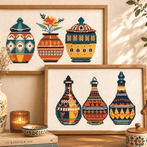 Puede incluir: Dos obras de arte en punto de cruz enmarcadas con diseños geométricos coloridos de jarrones y jarras. La obra superior muestra tres jarras con tapa y un jarrón con flores. La obra inferior muestra tres botellas decorativas.