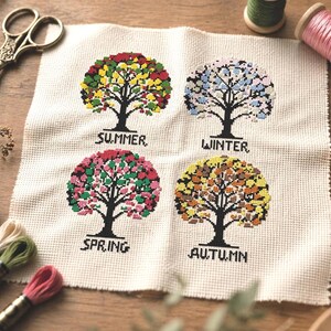 May include: A cross-stitch design on cream-colored fabric depicts four trees, each representing a season: Summer, Winter, Spring, and Autumn. The trees are in full bloom with colorful leaves. The word for each season is stitched below each tree.