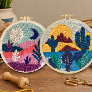 May include: Two round, framed textile art pieces with desert landscape designs. One features a moon and a crescent moon, the other a sun and cacti. The artwork is created with colorful yarn and embroidery, with a wooden punch needle and scissors in the foreground.