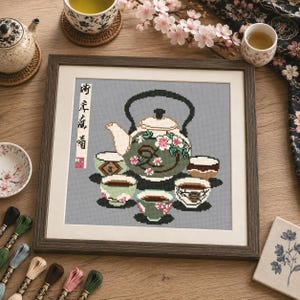 May include: A framed cross-stitch embroidery featuring a central green teapot decorated with pink cherry blossoms, surrounded by four small tea cups on a gray background. A vertical white banner with black Japanese calligraphy is on the left side.