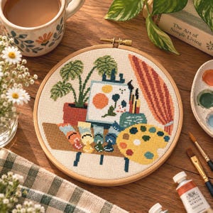 Op de afbeelding: Een ronde houten borduurring met een kruissteekontwerp van een kunstenaarswerkplek. Het patroon toont een monstera-plant in een pot, een canvas op een ezel met abstracte vormen, verftubes en een geel verfpalet.