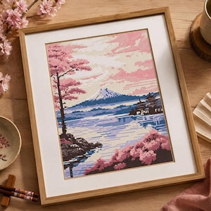 以下が含まれることがあります： 雪を頂いた山、湖、そしてピンク、青、白の色合いの桜が描かれたクロスステッチ刺繍の額装品。このアートワークは、白いマットボーダーが付いた明るい色の木製フレームに収められています。