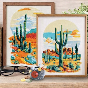 May include: Two framed cross-stitch artworks depict a desert landscape with cacti, mountains, and a setting sun. The artwork is in a wooden frame. A pair of glasses, a spool of thread, and buttons are on the shelf in front of the artwork.
