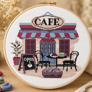 Peut inclure: Broderie au point de croix d'une scène de café dans un cercle en bois. Le café a un auvent rouge et bleu, une enseigne "CAFE" et un petit panneau "OPEN". Une table et des chaises sont installées à l'extérieur.