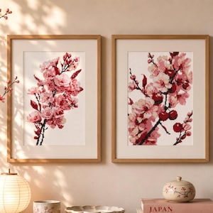 May include: Two framed floral art prints featuring pink cherry blossoms. The artwork is displayed on a light-colored wall, with a vase of cherry blossom branches and a paper lantern in the foreground. The frames are made of light-colored wood.
